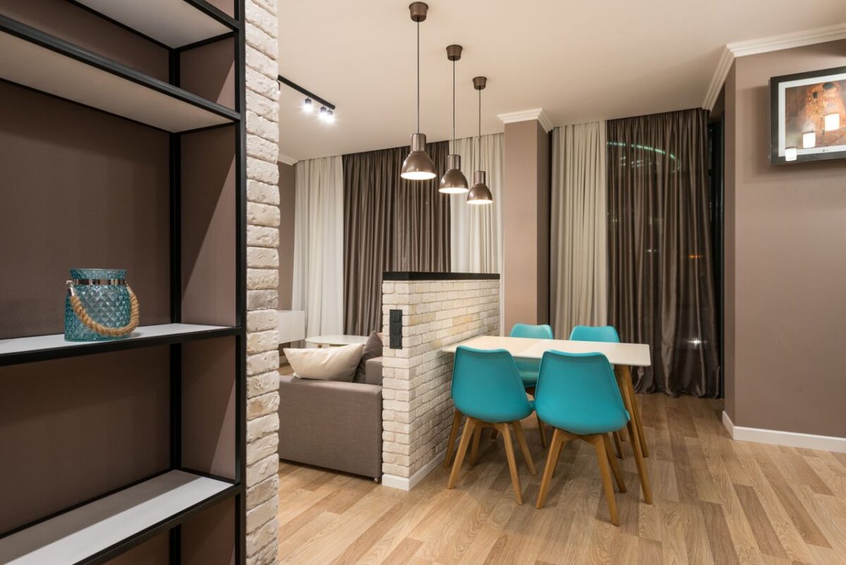 À propos Chairs at table placed near white brick partition in living room with couch in modern apartment with decoration on shelf
